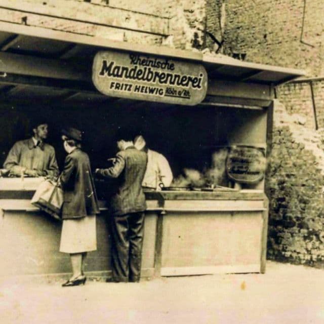 Historisches Sepia-Foto: Verkaufsstand der Rheinischen Mandelbrennerei Fritz Helwig in Köln, kurz nach dem Zweiten Weltkrieg, mit Fritz Helwig und Kunden am Tresen.